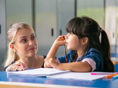 Serious female teacher discussing task with little pupil. Girl sitting at school desk, holding pencil and talking to tutor. Education or back to school concept